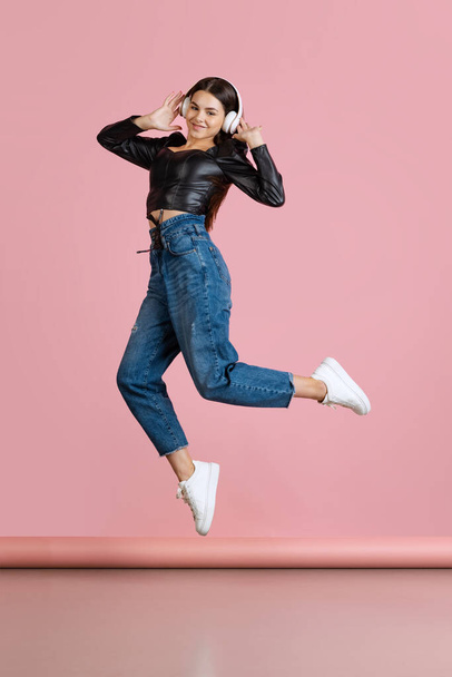 Full-length portrait of young beautiful Caucasian girl listening to music in headphones, jumping isolated over pink background - Fotografie, Obrázek