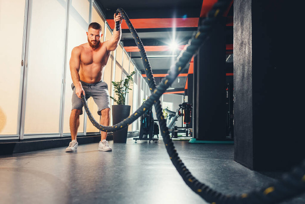 Athletic young man with battle rope doing exercise in fitness gym. Keeping fit with cross training. - Fotografie, Obrázek