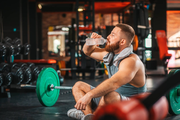 Молодой бородатый спортсмен тренируется в спортзале. Человек пьет бутылку воды. Концепция фитнеса и обучения. - Фото, изображение