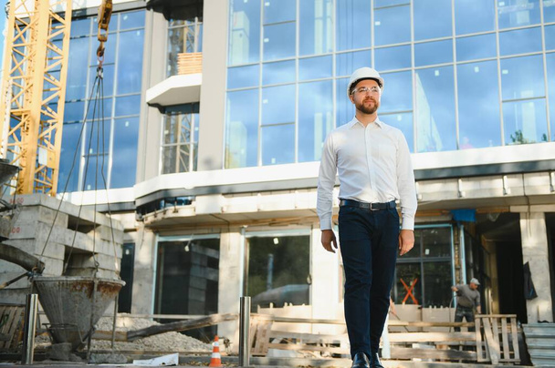 Successful engineer or architect, crane and building construction at backgrpound. Joyous businessman wearing helmet - 写真・画像
