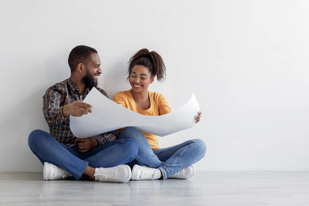 Happy funny young african american guy and lady look at blueprints at home, sit on floor over white wall background - Photo, Image