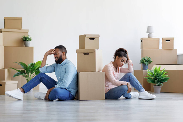 Tired upset young black husband and wife among cardboard boxes with things and plants in empty room - Photo, Image