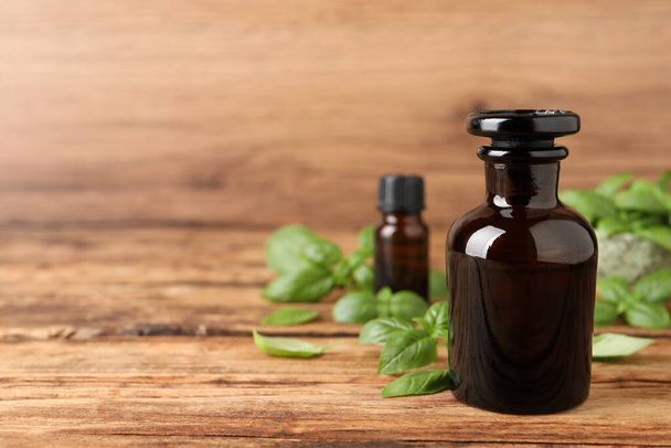 Glass bottles of basil essential oil and leaves on wooden table, space for text - Fotó, kép