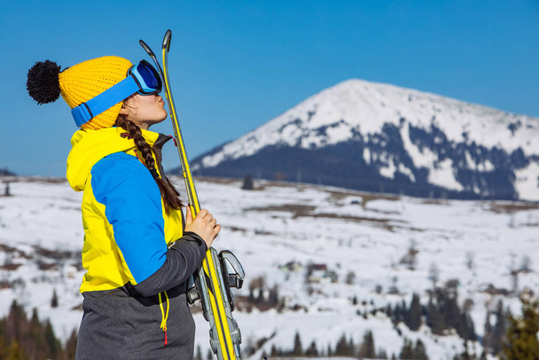 joven sonriente bonita mujer sosteniendo esquí. montañas en el fondo. viajes de invierno. espacio de copia
 - Foto, Imagen