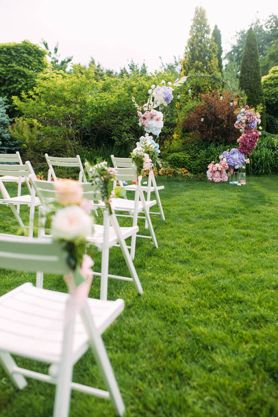 Весільна церемонія в прекрасному ландшафтному саду серед газону зі стильною весільною аркою прикрашена свіжими квітами і білими стільцями поруч
. - Фото, зображення