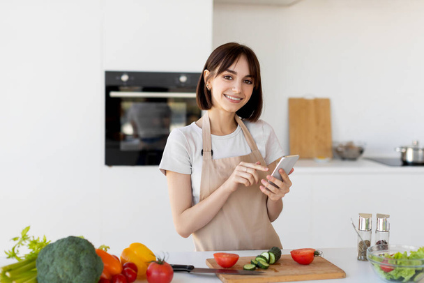 Glückliche Millennial-Frau nutzt Smartphone und sucht nach neuen Rezepten, steht in der Küche und lächelt in die Kamera - Foto, Bild