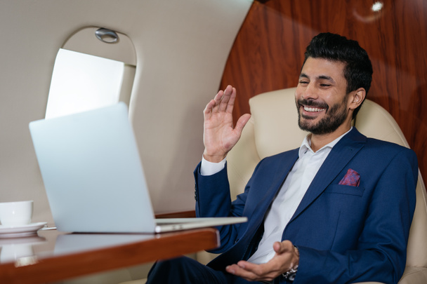 Schöner lächelnder arabischer Geschäftsmann mit Laptop und Videoanruf im Luxus-Privatjet. Online-Meeting, Videokonferenzkonzept - Foto, Bild