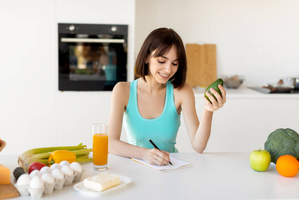 Koncepcja diety keto. Happy fit kobieta pisząc codzienną dietę w notatniku, siedzi w pobliżu różnych produktów przy stole kuchennym - Zdjęcie, obraz