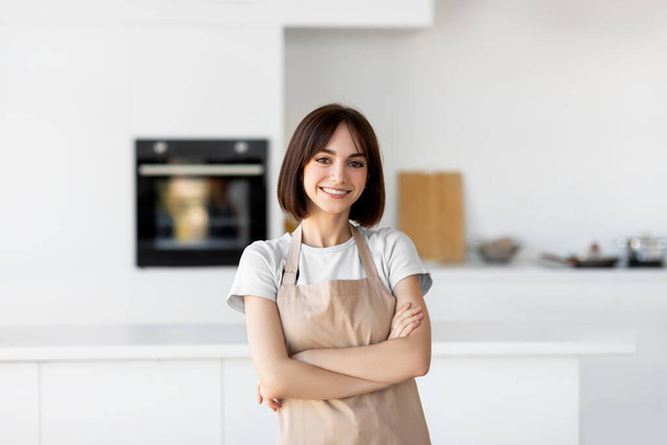 Fiatal fehér hölgy kötényben áll összecsukott karokkal, néz és mosolyog a kamerába, pózol a konyhában - Fotó, kép