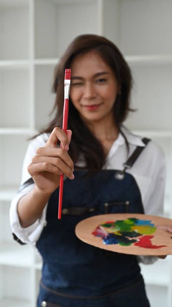 Portrait female artist with brush and paint palette standing in her art studio. - Photo, Image