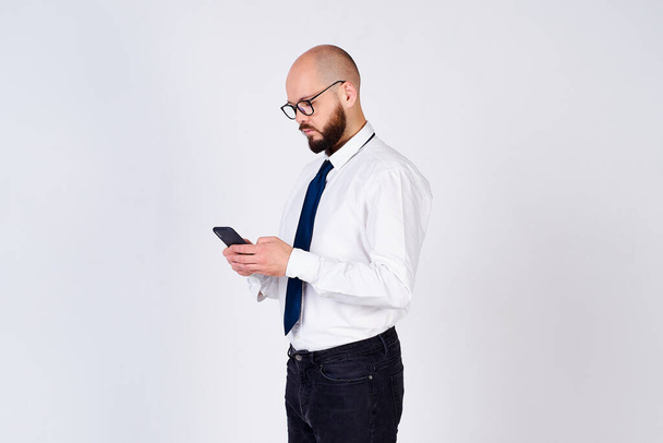 Serious handsome mature man, businessman, in a white shirt, tie, wears glasses, stands sideways and uses a mobile phone, is isolated on a white background. - Photo, image