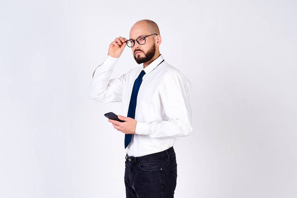 Confident mature man, bald with a beard, businessman, in a white shirt, tie, wears glasses, looks at the camera and uses a mobile phone, is isolated on a white background. - 写真・画像