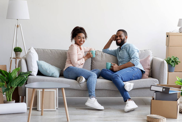 若いアフリカ系アメリカ人の男性と女性の笑顔はソファでリラックスし、段ボール箱付きの部屋でコーヒーを飲む - 写真・画像