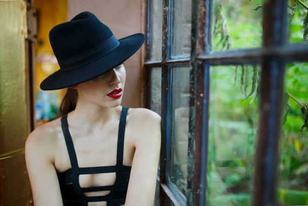 Μυστήριο νεαρή γυναίκα σε μαύρο καπέλο και μεγάλη μακιγιάζ με κόκκινα χείλη, κάθεται κοντά στο παράθυρο, κοιτάζοντας μακριά. - Φωτογραφία, εικόνα