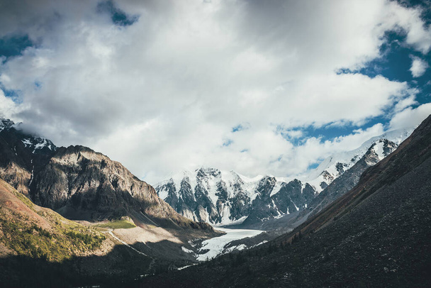 Prachtig berglandschap met gletsjertong en grote besneeuwde bergen in het zonlicht in de bewolkte lucht. Scenic alpine landschap met gletsjertong en hoge bergen met gletsjers in lage wolken. - Foto, afbeelding