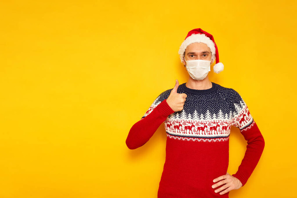 man in Christmas sweater with reindeer, medical mask, New Years hat, a Santa Claus hat, gives a thumbs up, likes, OK sign. isolated yellow background with space for text. concept holiday, gifts, sale - Foto, Imagem