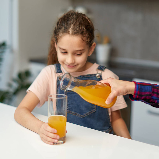 Κοντινό πορτρέτο ενός χεριού ρίχνει χαριτωμένο κοριτσάκι χυμό πορτοκάλι για πρωινό - Φωτογραφία, εικόνα