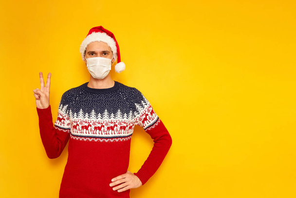 man in New Years sweater with reindeer, medical mask on his face, Santa Claus hat shows sign of V, peace, victory. isolated yellow background with space for text. concept new year, Christmas, holiday - Zdjęcie, obraz