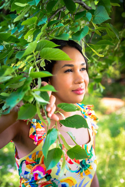 A beautiful mixed race African American woman in yellow floral dress standing in the forest on a sunny summer day parts a tree branch covered in leaves with her hand. - Фото, зображення