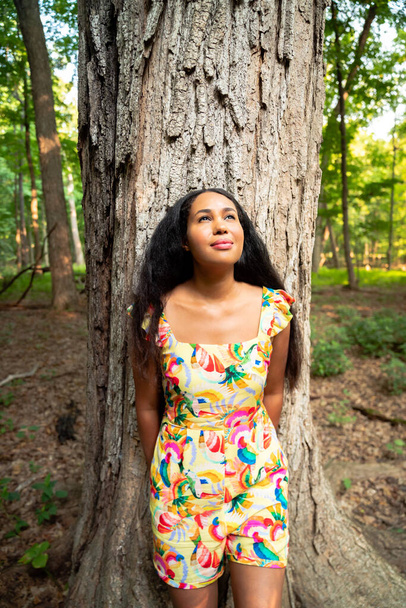 Belo retrato de uma mulher afroamericana de raça mista feliz vestindo um vestido floral amarelo encostado a um grande tronco de árvore coberto de casca áspera em uma floresta e olha para a distância. - Foto, Imagem