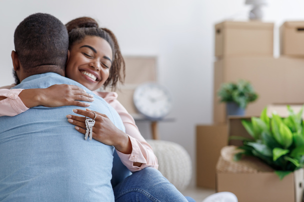 Satisfied young black family hugging, hold key and enjoy buying real estate in room with cardboard boxes and plants - Photo, Image