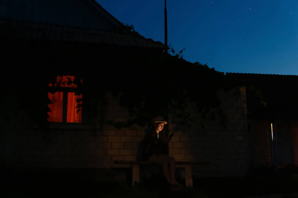 Дефокусна жінка використовує смартфон, сидячи на лавці вночі біля будинку. Зовнішній портрет тисячоліття, використовуючи свій мобільний телефон вночі. Помаранчеве світло у вікні. Зірки в літньому нічному небі. За межами фокусу
. - Фото, зображення