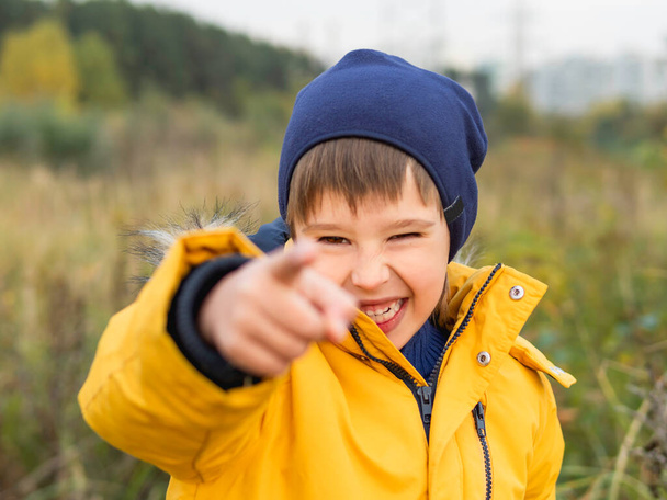 Smiling boy in yellow jacket points his finger at camera. Laughing kid walks in park. Leisure activity outdoors. Bright clothes for fall season.  - Photo, image