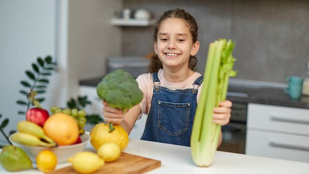 Smiling pretty little girl stand near table in the kitchen, holding green broccoli and celery in hands. - Photo, image