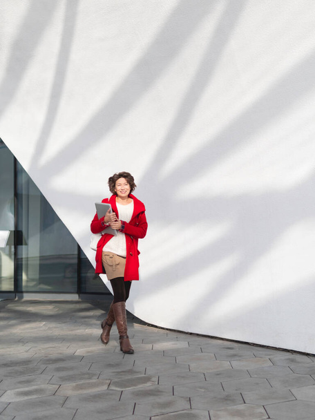 Curly woman in red coat is walking by white wall. Smiling student with laptop. Freelancer with portable device for work. Modern lifestyle. - Фото, зображення