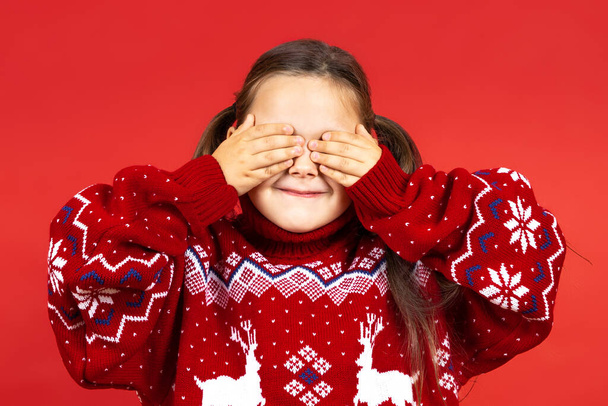 közelkép portré mosolygós, bájos lány piros karácsonyi pulóver rénszarvas eltakarja a szemét a kezét, és várja az ajándékot, elszigetelt piros háttér - Fotó, kép
