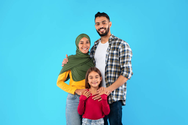 Modern Muslim Family Of Three Hugging Standing Over Blue Background - Fotó, kép