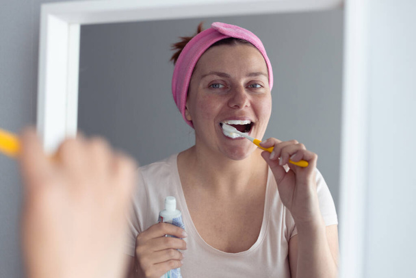 Fiatal, gyönyörű nő fogkefével a tükör mellett a fürdőszobában. Szájhigiénia és fogápolás - Fotó, kép