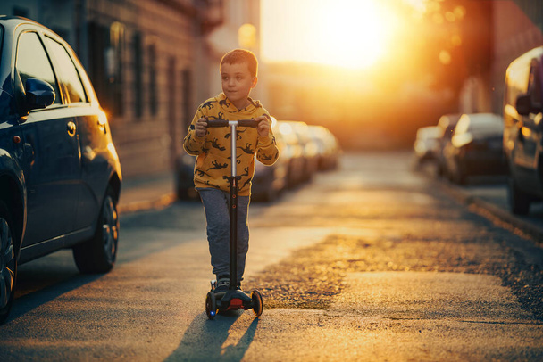 アクティブ子供男の子に乗る彼のスクーターで街で日没時. - 写真・画像
