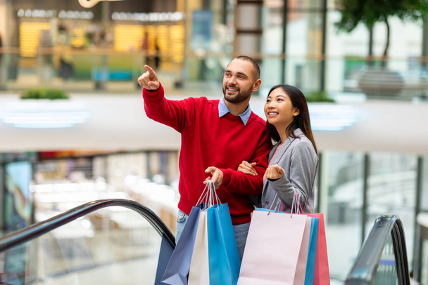 Feliz pareja diversa con bolsas de regalo utilizando escaleras móviles, ir de compras juntos en el centro comercial de la ciudad - Foto, Imagen