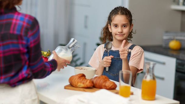Hymyilevä pieni tyttö, jolla on kiharat hiukset, näyttää peukalot, joilla on terve aamiainen yhdessä äidin kanssa keittiössä.. - Valokuva, kuva