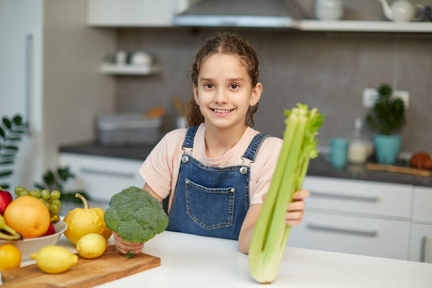 Smiling little girl stand near table in the kitchen, holding green broccoli and celery in hands, looking at camera. - Photo, image