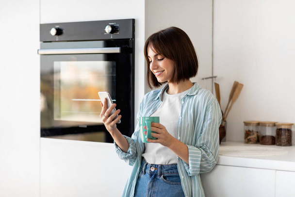 Social networks and chat in morning. Happy young lady drinking coffee and using smartphone, standing in kitchen - Foto, Bild
