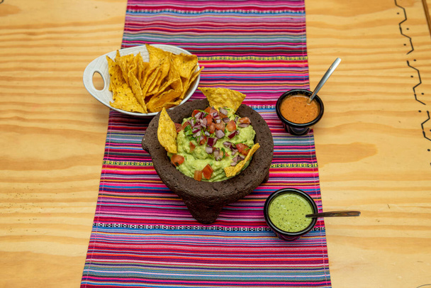 Mexican volcanic stone molcajete filled with guacamole with corn chips, cheese sauce and hot sauce with pico de gallo - Photo, Image