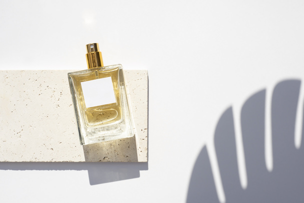 Transparent bottle of perfume with white label on stone plate on a white background. Fragrance presentation with daylight. Trending concept in natural materials with palm leaf shadows. - Photo, Image
