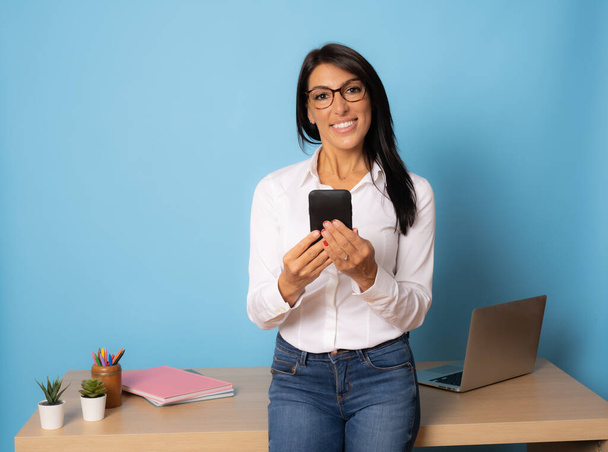 Vonzó mosolygós üzletasszony támaszkodik az asztalra segítségével okostelefon felett kék háttér - Fotó, kép