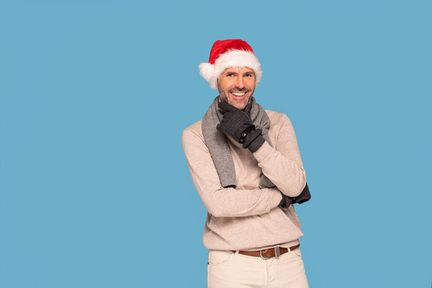 Handsome smiling man wearing winter clothes and red Santa Claus hat. Blue pastel studio background.  A lot of copy space. - Fotografie, Obrázek