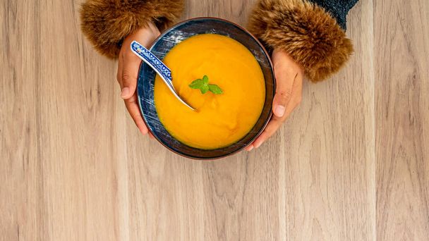 Ręce trzymające zupę dyniową na stole. Kobiece ręce trzymające miskę zupy dyniowej z liściem mięty. Jedzenie sezonowe. - Zdjęcie, obraz
