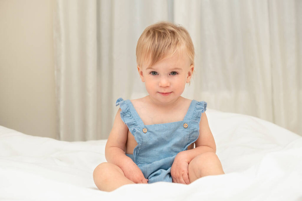 Cute pretty beautiful serious blonde caucasian baby girl abot 1-2 years old, wearing blue body suit,looking at camera,infant,sitting on white blanket bed in home cozy bedroom.Copy space. - Foto, imagen