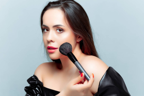 a beautiful young brunette is applying tonal powder with a makeup brush on her face - Photo, Image