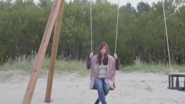 Authentic girl riding a swing, she is wearing a burgundy jacket and blue jeans, she is on the beach by the baltic sea, autumn day - Footage, Video