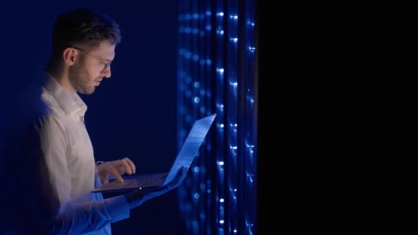 Arc portré egy férfiról, aki a szerverszobában dolgozik laptoppal. Egy technikus ellenőrzi a szerver szobát. Kaukázusi mérnök szürke munkáskabátban - Felvétel, videó