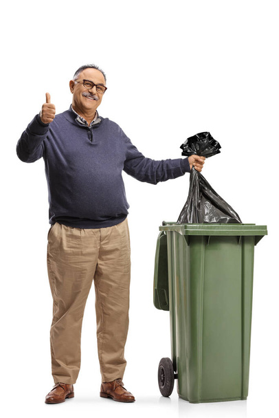 Mature man throwing a plastic bag in a dustbin and gesturing a thumb up sign isolated on white background - Photo, Image
