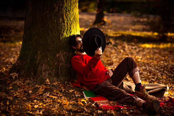 a brutal young man with a beard, wearing a black hat and a red sweater, in the afternoon in the park under an autumn tree - Foto, Imagen