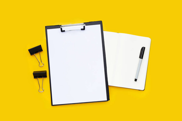 Leeres A4-Papier in schwarzem Klemmbrett, schwarze Büroklammern mit Notizbuch und Stift auf gelbem Hintergrund. - Foto, Bild