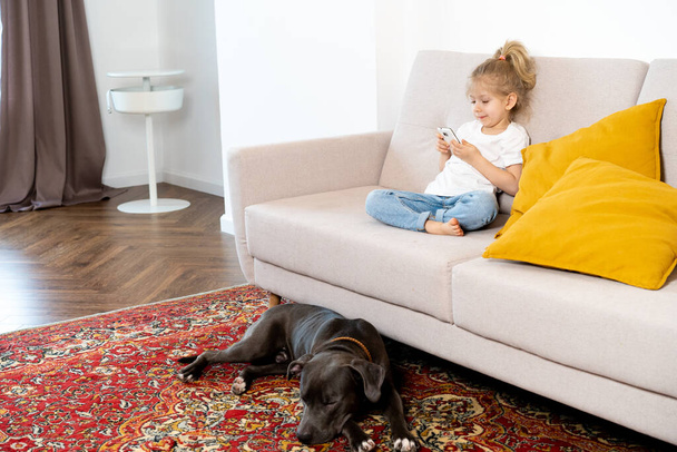 ein kleines blondes Mädchen zu Hause auf dem Sofa mit Handy und großem Hund, Kindern und Gadgets. - Foto, Bild
